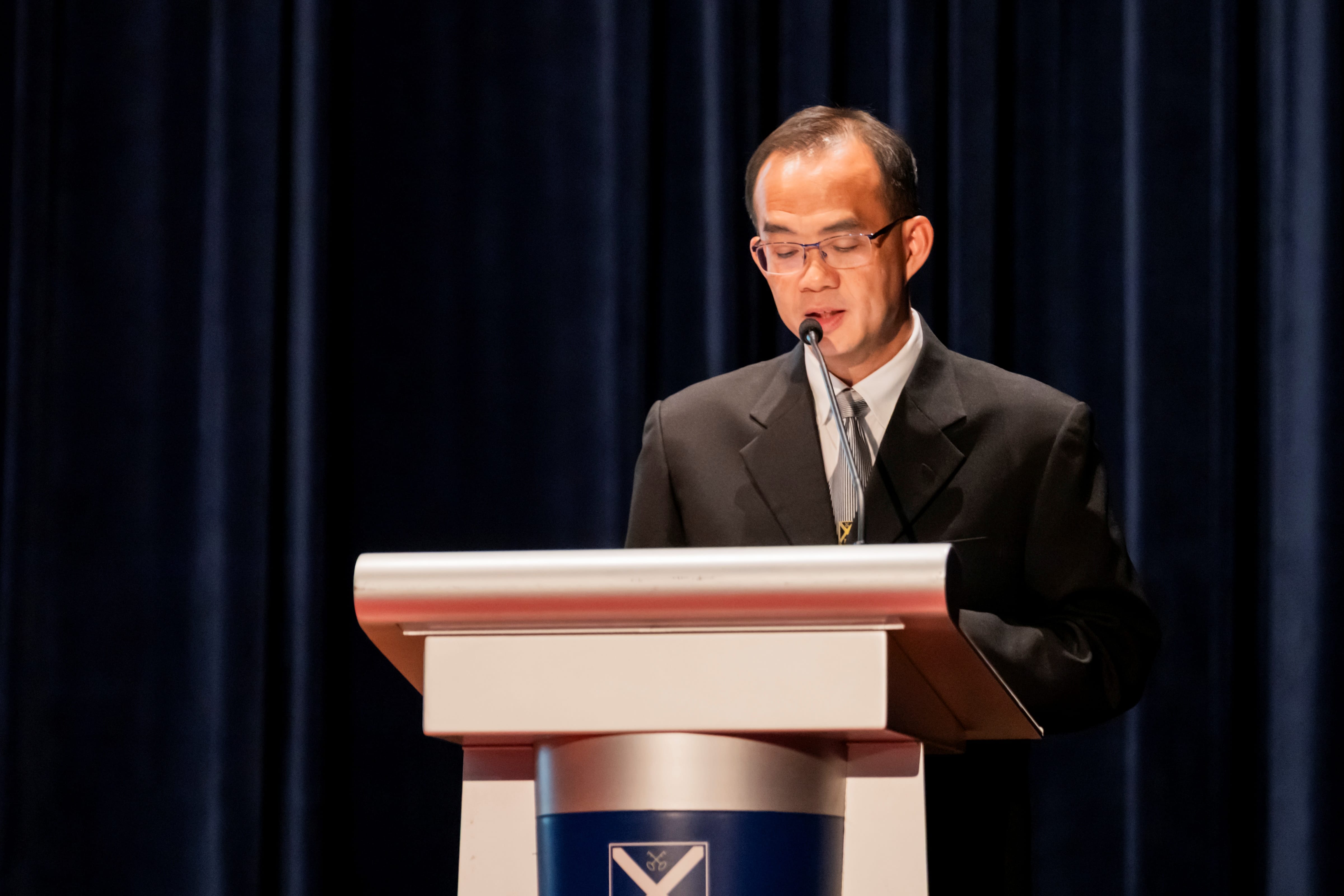 Principal, Mr Tham Kine Thong, in a dark suit speaking at a podium against a dark curtain backdrop, delivering the Principal's Report.